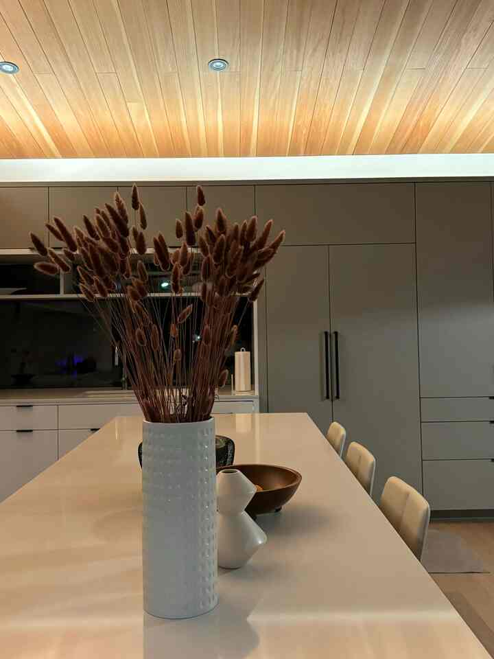 Wood tone ceiling and minimalist modern kitchen featuring a long table with bar stools and a vase centerpiece