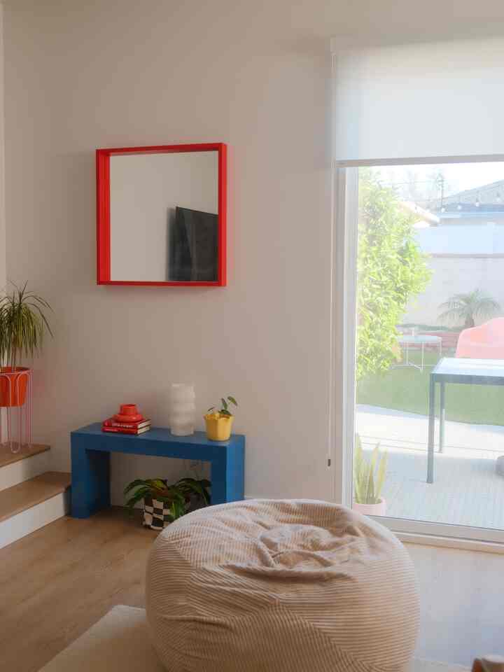 Bright ivory-toned living room featuring a comfortable bean bag at center, red-framed mirror, blue bench with plants on the left creating a cozy atmosphere