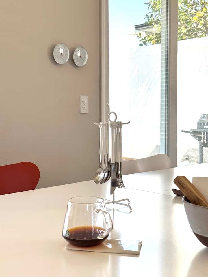 Bright beige wall and white dining table featuring a transparent glass mug of coffee and white-handled cutlery set in a clean home cafe space