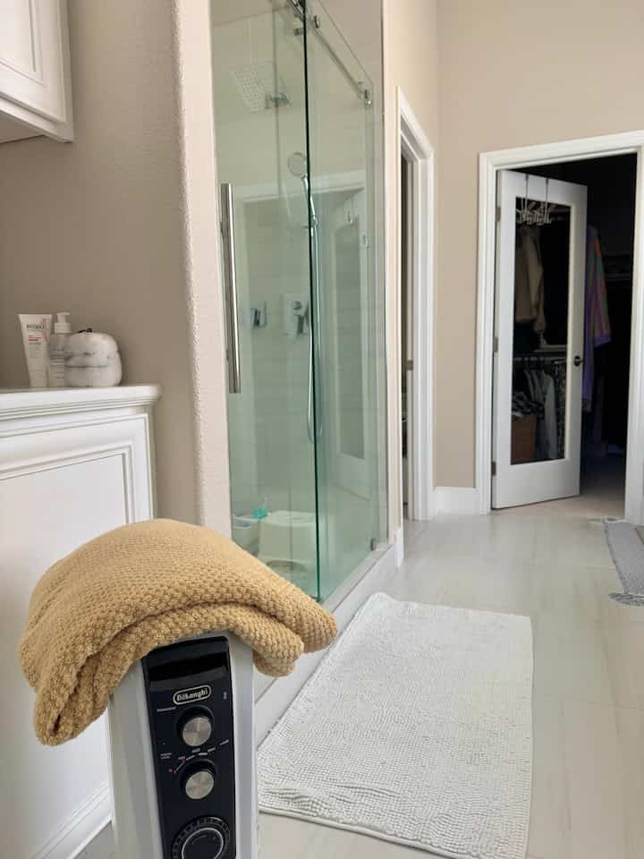 White and beige toned bathroom featuring a glass shower cabin and a portable heater with a beige towel on top, creating a warm atmosphere