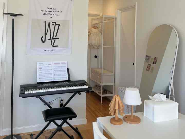 A simple small room with white and wood tones featuring an electronic piano, large mirror, floor lamp, and storage shelf