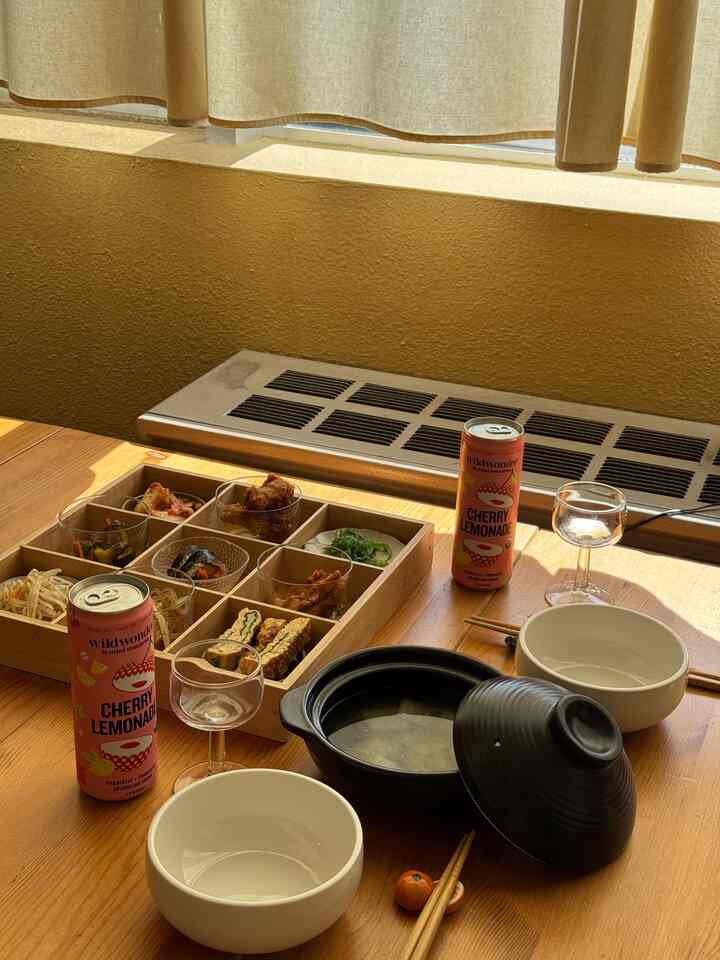 Cozy kitchen dining space with warm orange wall and natural wood table, featuring Korean lunch box and beverages