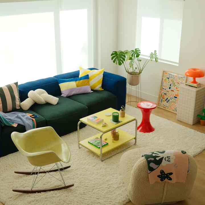 Modern living room with blue and green sofa, yellow coffee table on white shag rug, bright natural lighting and plant accents