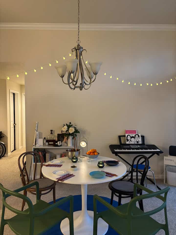 White and beige toned dining room featuring a round dining table, four dining chairs, a central chandelier, and string lights creating a cozy home party atmosphere