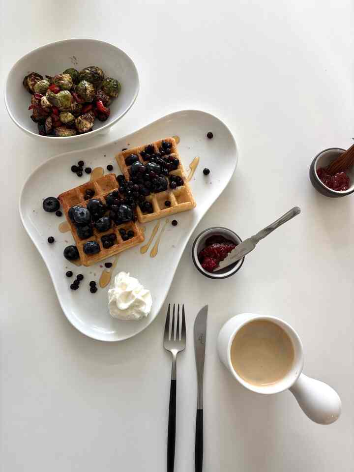 White table featuring waffles with blueberries, cutlery, and coffee mug in a neat home cafe brunch setup