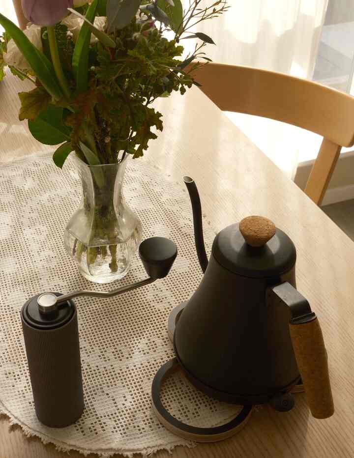 Natural-colored kitchen table featuring a black electric kettle, coffee grinder, and transparent glass vase creating a cozy home cafe atmosphere