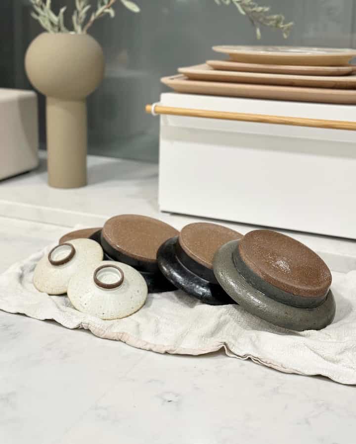 White and beige toned kitchen counter featuring handmade ceramic plates and bowls neatly arranged on a towel, natural and cozy atmosphere