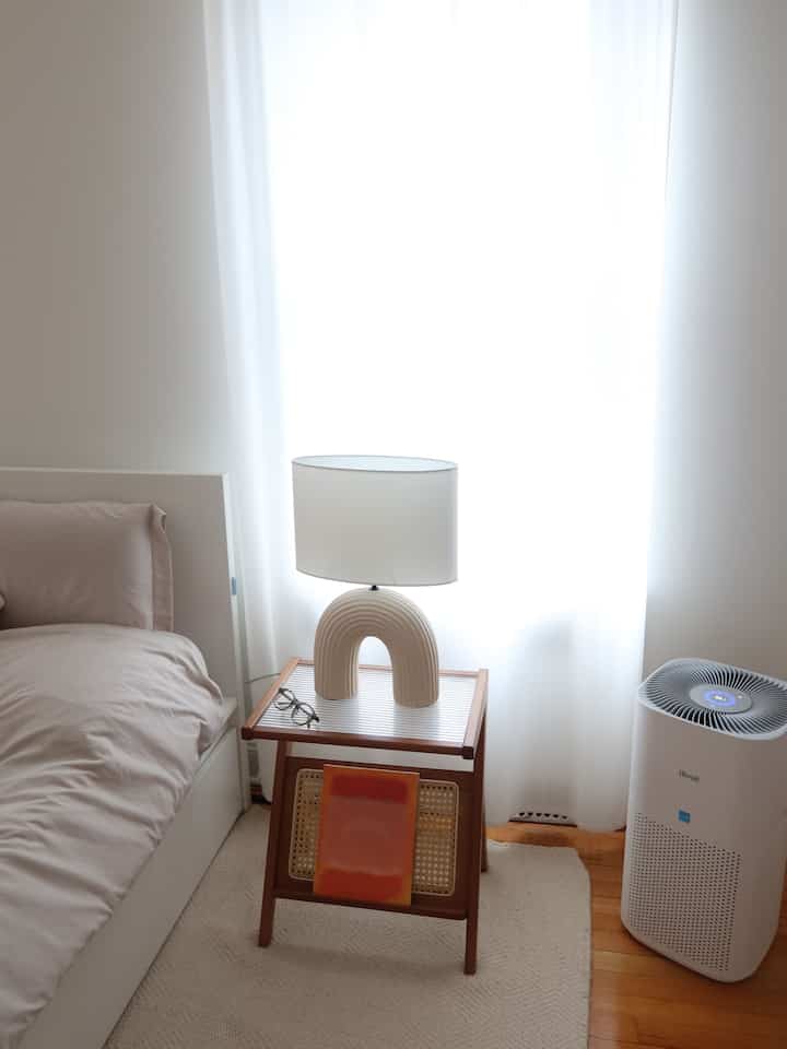 White-tone bedroom featuring white curtains and carpet, wooden side table with unique lamp creating a simple atmosphere