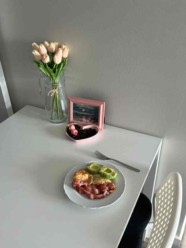 White-toned dining room featuring a table with avocado, bacon, eggs, a flower vase, and small decor, creating a clean atmosphere