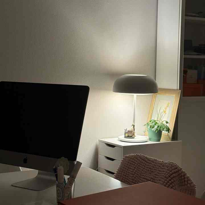 White-toned home office space featuring an iMac on a desk with a table lamp and a plant, creating a neat and modern atmosphere
