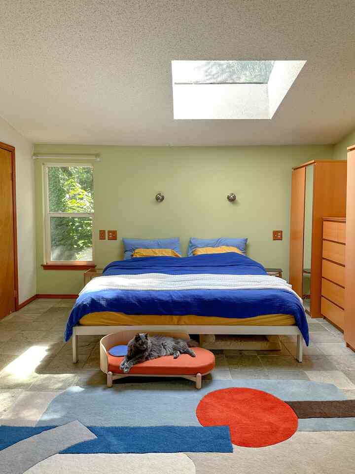 Natural-toned bedroom with a centrally placed bed covered in blue duvet and a cat resting on an orange pet bed in front