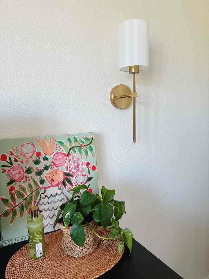 Bright white wall with gold wall sconce in bedroom corner featuring green plant and handmade rattan placemat creating natural vibe