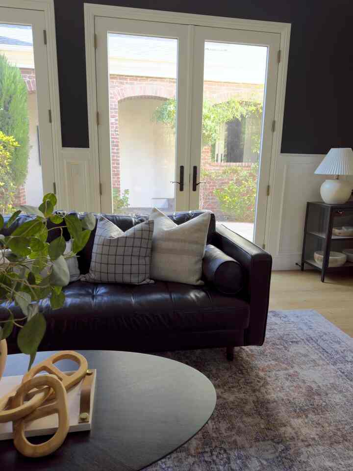 Bright white living room with large windows, featuring a dark brown leather sofa, pillows, rug, and table lamp in a natural modern style