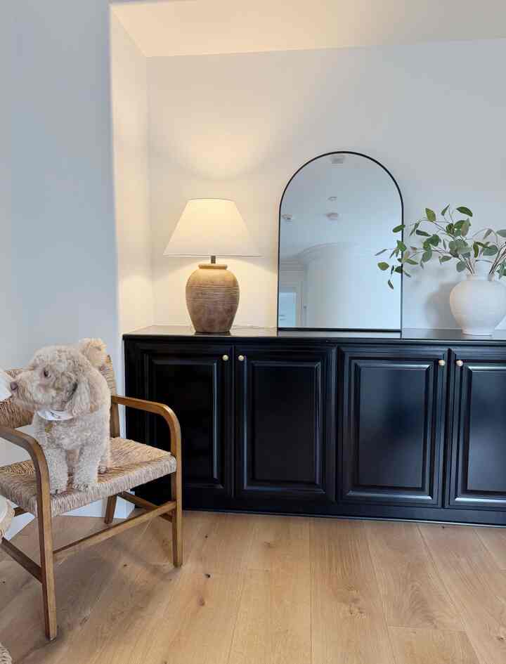 Natural wood-tone entrance space featuring a wooden chair, arched black mirror, black cabinet, and a table lamp with modern ambiance