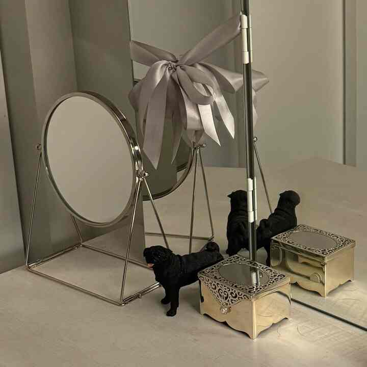 A clean silver-toned vanity table featuring two mirrors, a black dog figurine, and an ornate decorative box.