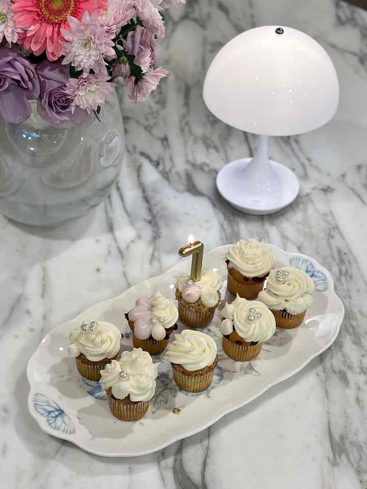 White-toned kitchen table featuring cupcakes on a decorative plate, a white table lamp, and a vase with flowers, creating a cozy stylish atmosphere