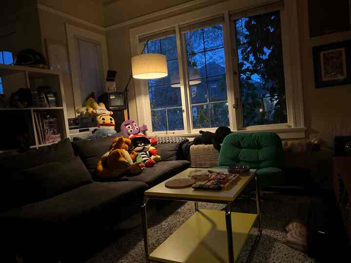 Dimly lit living room with black sofa, green armchair, yellow side table, and colorful plush toys near large window