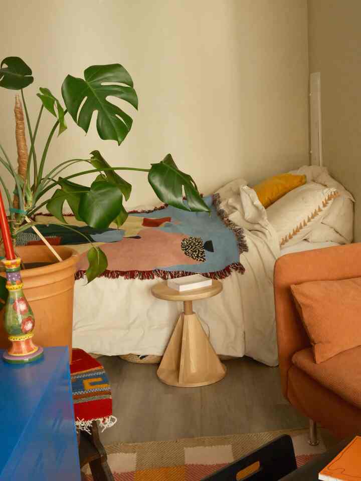 Beige and orange toned studio apartment featuring a bed, orange sofa, wooden stool, and large leafy plant creating a cozy atmosphere