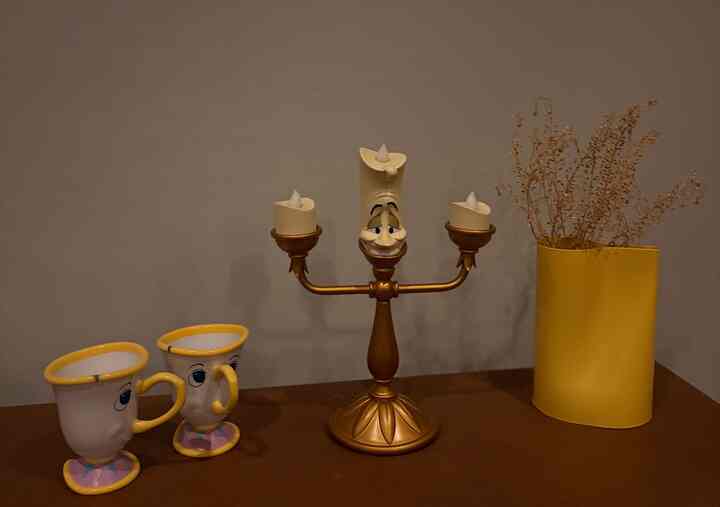 Gold and yellow toned decorative candelabra, yellow vase, and character-themed mugs arranged on a cozy decorative surface