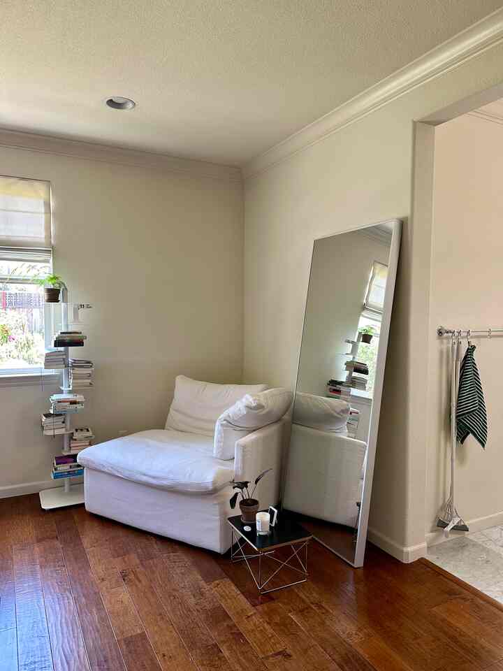 A cozy bedroom corner with white and wood tones, featuring an armchair, floor mirror, and bookshelf