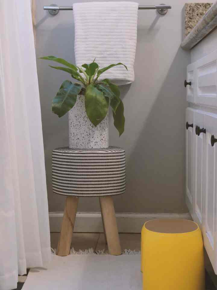 Bathroom space with white and beige tones featuring a striped stool topped with a plant, complemented by white towel and curtain in a clean setting