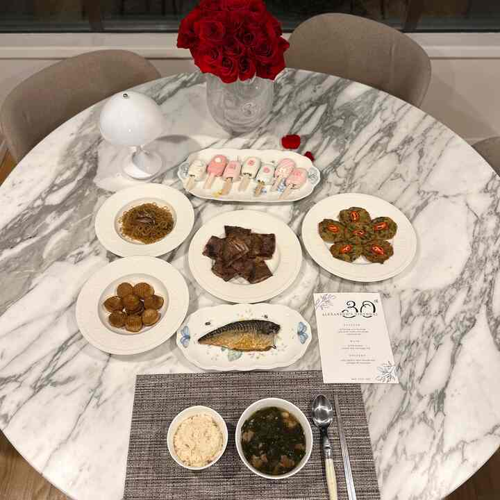 White and marble-toned dining room featuring a marble dining table centered with birthday floral arrangement and assorted Korean dishes neatly set