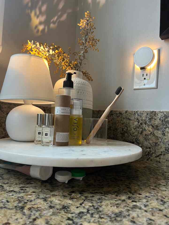 White-toned bathroom vanity featuring a marble Lazy Susan with a table lamp, body oil, and a toothbrush arranged neatly