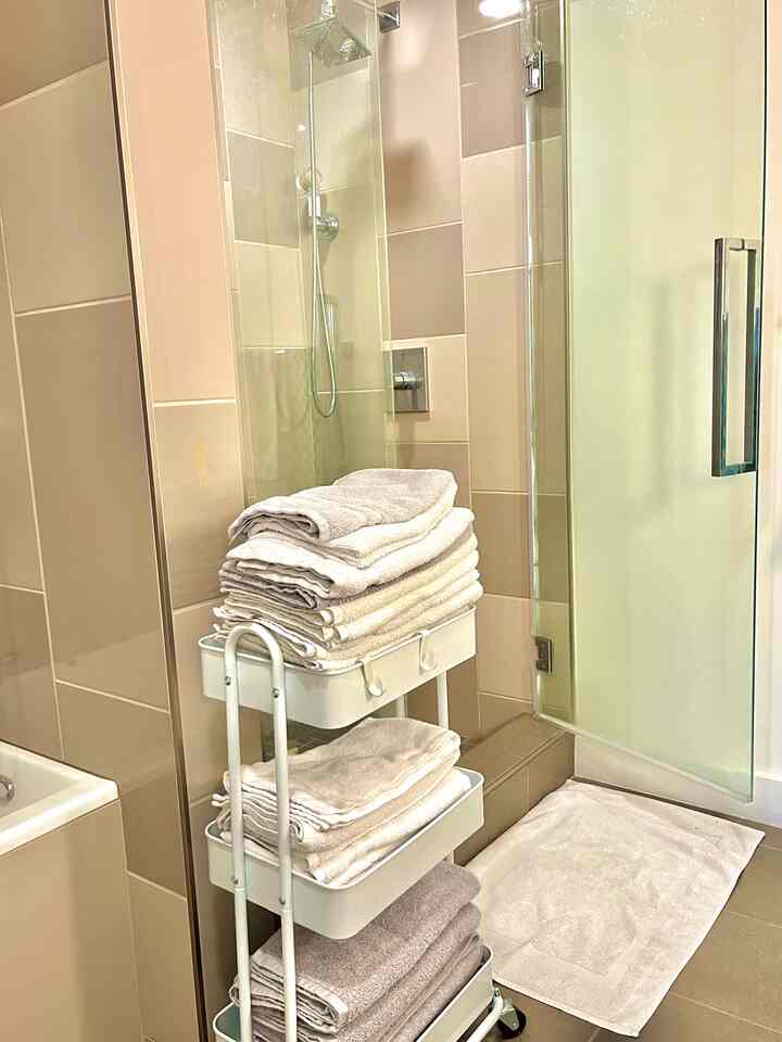 White and beige toned bathroom featuring stacked towels on a metal cart and a bath mat, creating a clean atmosphere