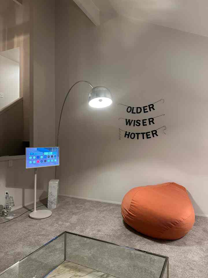 Living room corner with white walls and gray carpet floor, featuring an orange bean bag and silver arc floor lamp creating cozy atmosphere