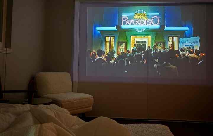 Cozy bedroom with white blankets and brown tones, featuring a movie projection on the wall creating a warm home cinema atmosphere