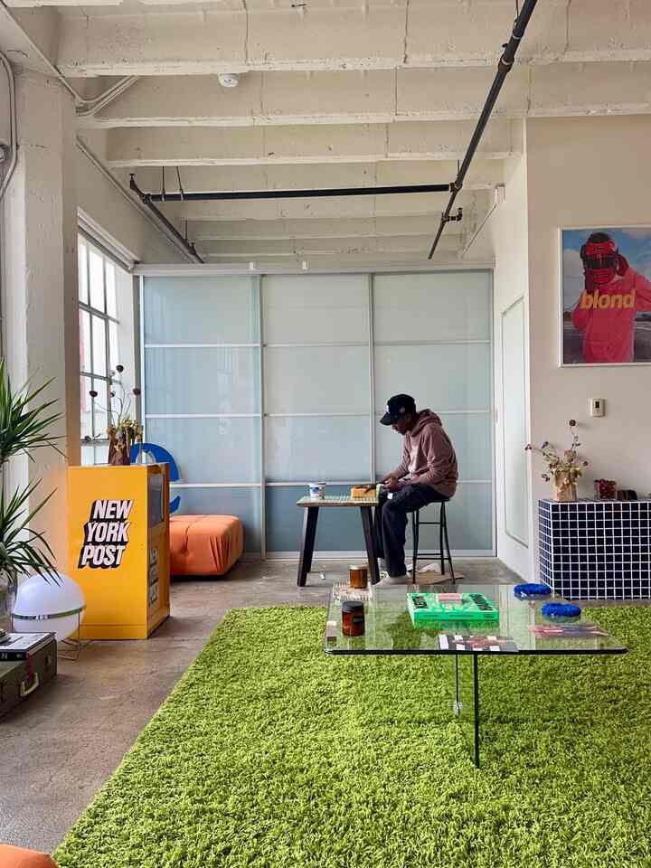 Natural and simple living room with a bright green rug, a person working remotely seated on a stool near window