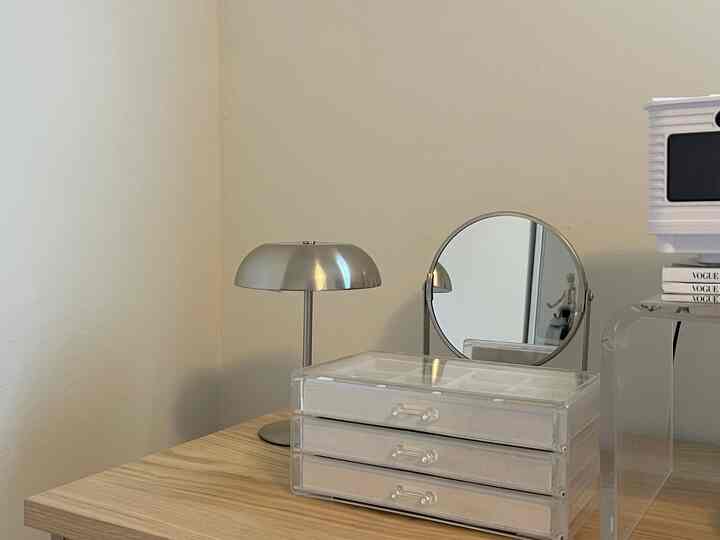 White and silver toned table scene featuring a vanity setup with a jewelry box, round mirror, and table lamp in a clean space