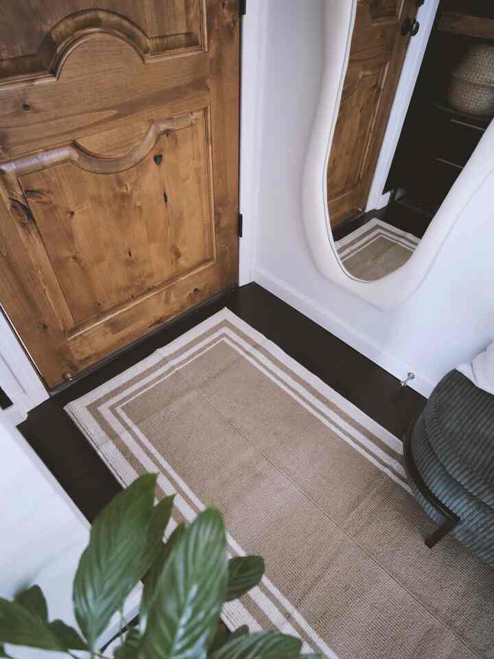 A small entrance with beige and brown tones featuring a wooden door, long rug, and wall-mounted mirror in a natural setting