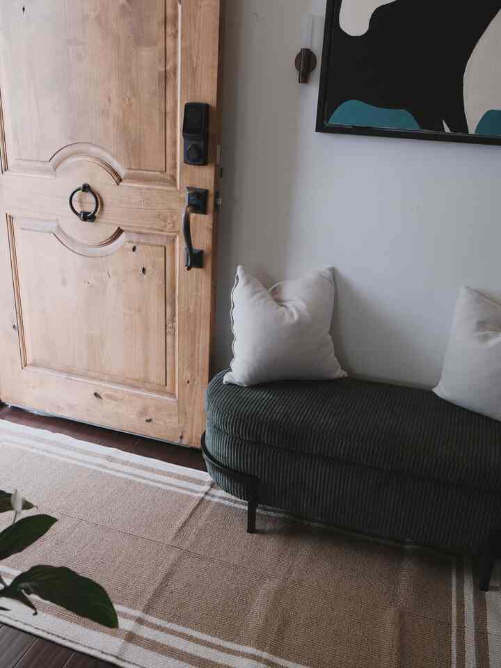 Natural-toned entrance featuring a brown wooden door, beige rug, and dark green ottoman with cushions