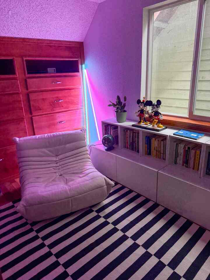 White bookshelf and navy striped rug in a home office with pink neon lighting and a lounge chair creating a cozy atmosphere