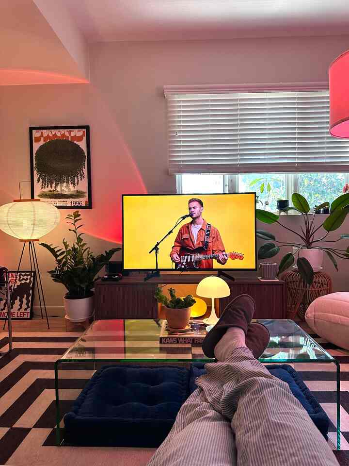 Modern living room with red accent lighting and plants, featuring a glass coffee table and centrally placed TV for a cozy atmosphere