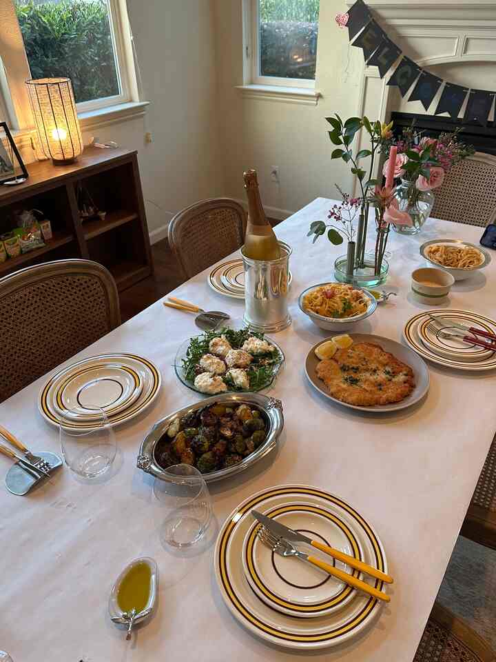 White tablecloth-covered dining room with brown dining table and chairs, cozy space set for a home party with food and flower vases