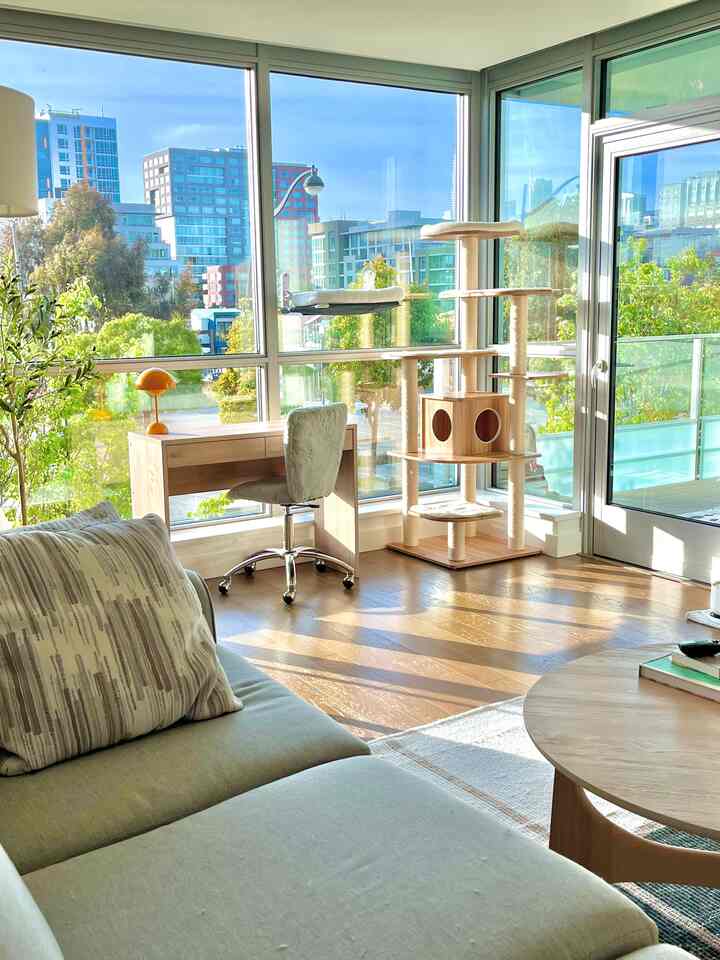 Bright wood-tone living room with large windows featuring sofa, desk, and cat tower creating a natural atmosphere