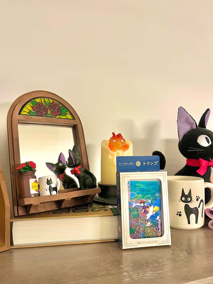 A natural-toned bookshelf corner featuring Ghibli character collectibles, mugs, and a mirror creating a cozy nook