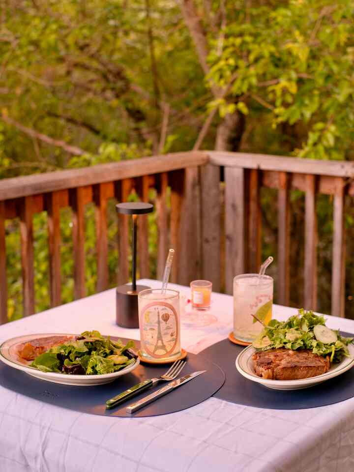 White-tablecloth outdoor dining table on brown wood veranda surrounded by green leafy trees, set with plates and cutlery