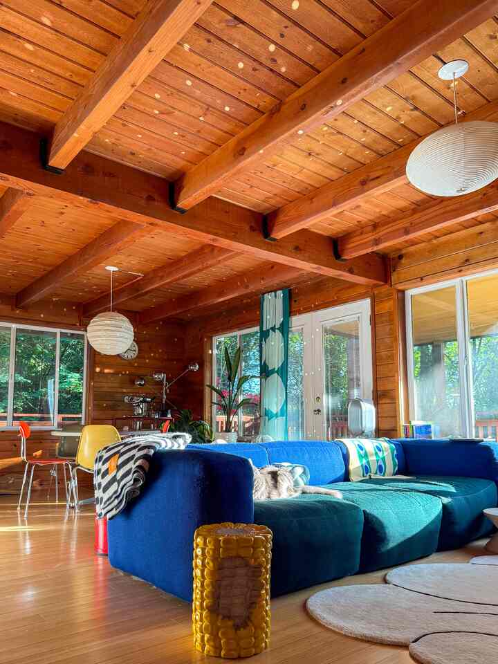 Natural wood-paneled living room featuring a blue sectional sofa and a yellow corn-shaped stool, creating a warm atmosphere