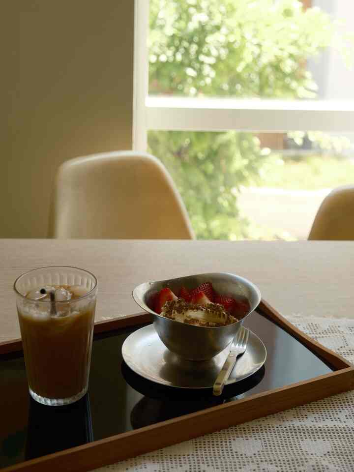 Beige-toned dining room with wood tone table featuring a tray holding iced coffee and strawberry dessert near a bright window