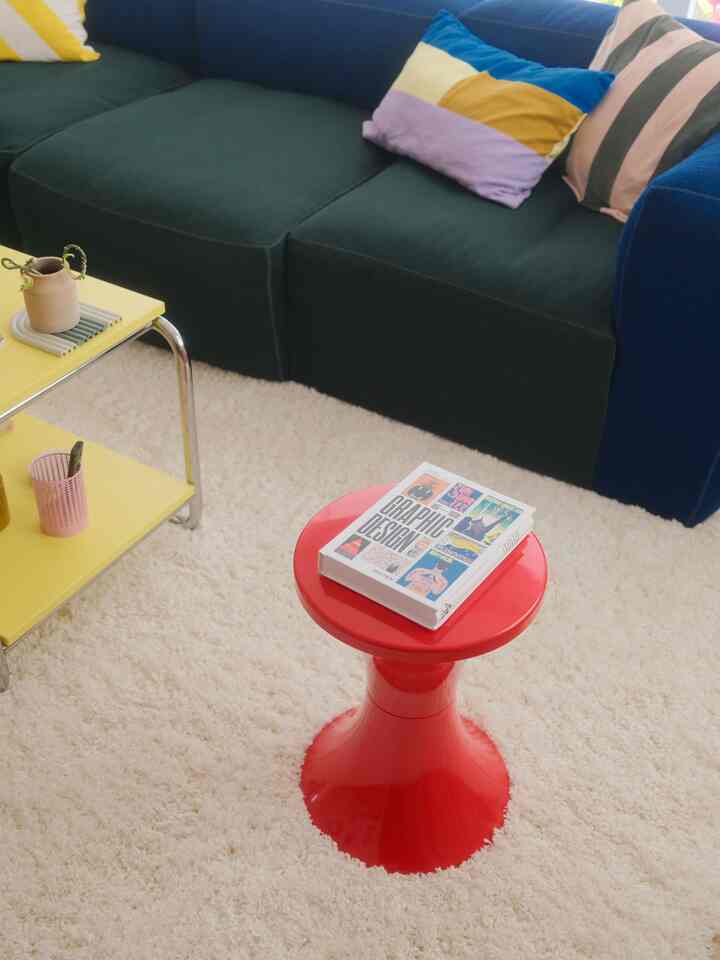 Living room with dark blue sofa, red stool, yellow side table on cream rug, creating a cozy and colorful vibe