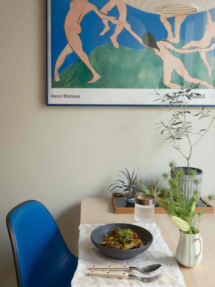 Natural-toned dining room featuring a wooden table, blue dining chair, a bowl of food on a white cloth, and green plants creating a cozy atmosphere