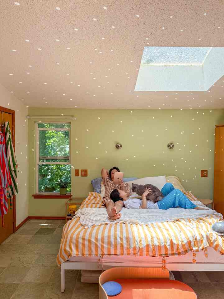 Natural light green bedroom featuring a large bed with orange striped duvet cover, two people relaxing with a cat amid a cozy atmosphere