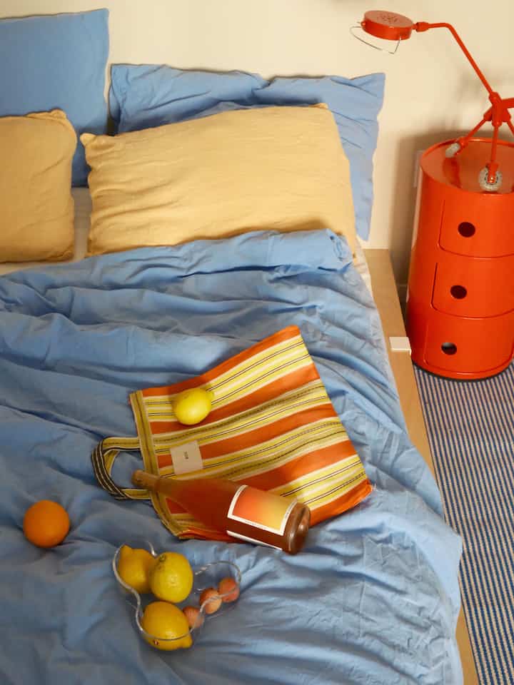 Cozy bedroom with soft blue and yellow pillows and an orange bedside drawer, featuring a casual bed setup
