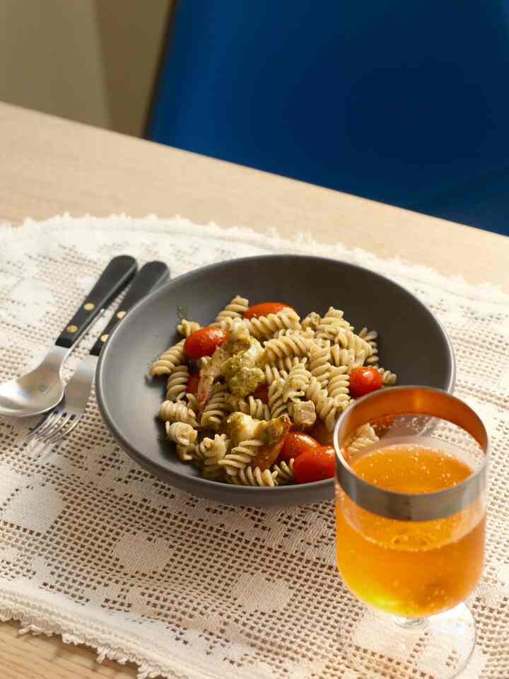 Dining table with wood tone featuring bowl of pasta, cutlery and orange drink on white crochet table runner evoking home cafe vibe