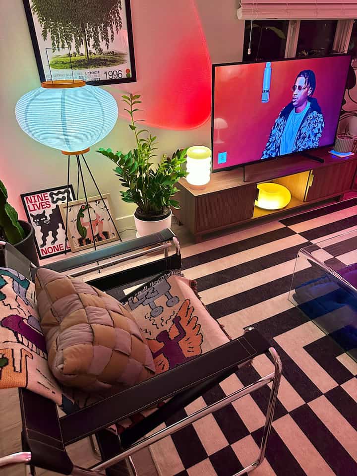 Modern living room with a blue glowing floor lamp, natural wood TV stand, striped rug, and transparent chair creating a cozy atmosphere