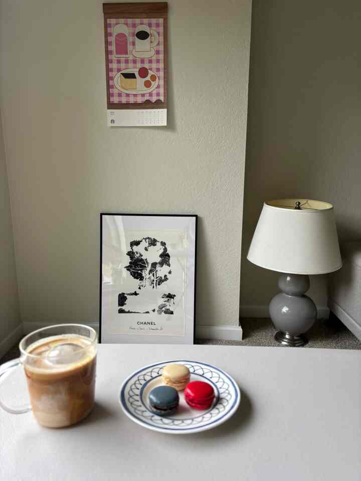 Bright white-toned corner space featuring a coffee table with coffee and macarons plate, table lamp and framed art creating cozy cafe style interior