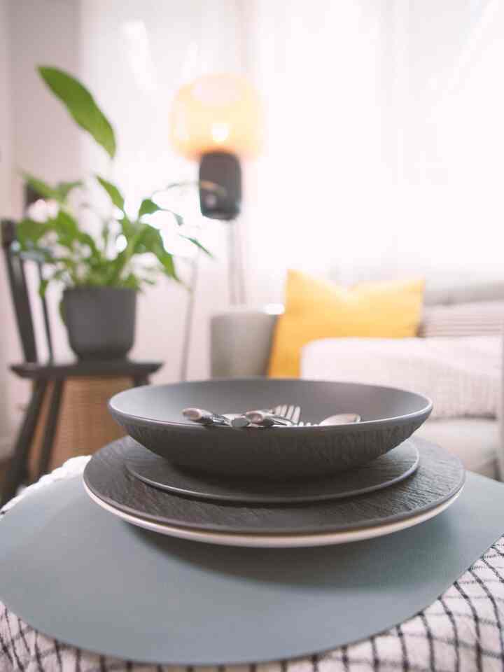 Bright-toned living room featuring stacked black plates and cutlery in foreground with sofa and yellow cushion in background creating a warm atmosphere